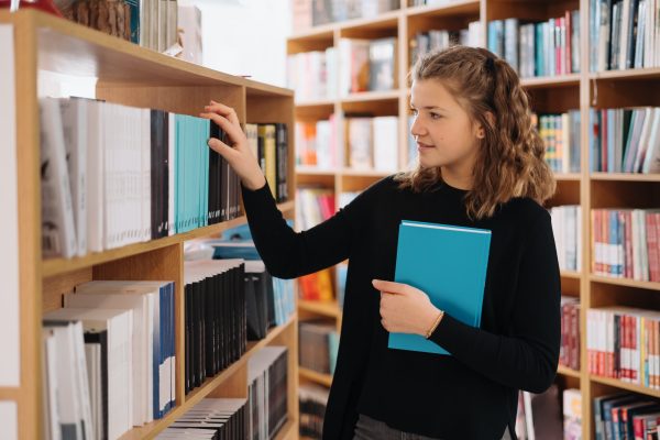 Teenager girl picking a book on a library shelf